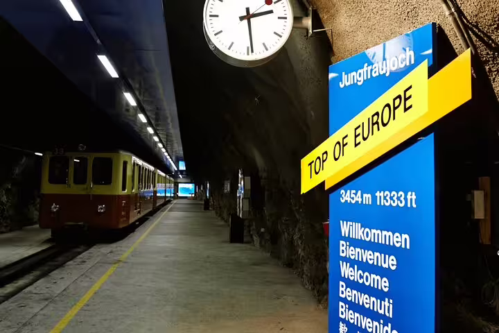 Jungfraujoch railway station platform with a train, clock, and multilingual welcome sign at 3454m elevation.