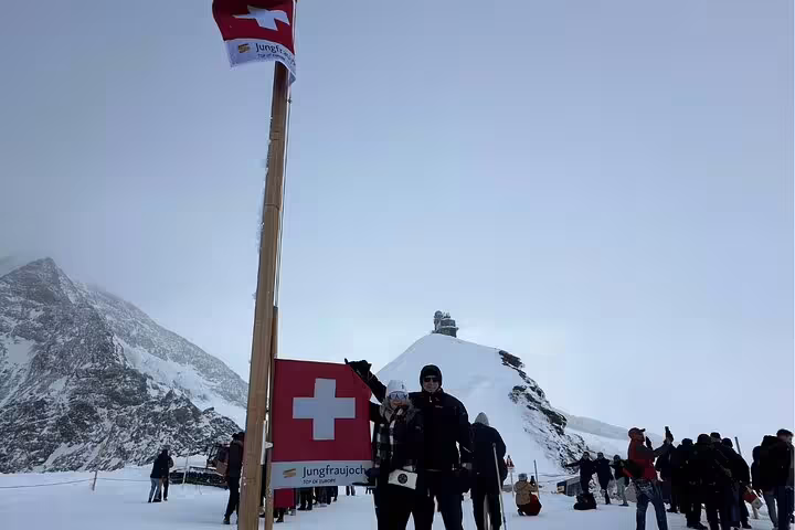 Tourists posing by Swiss flag at Jungfraujoch summit, capturing iconic Alpine experience on private day trip.