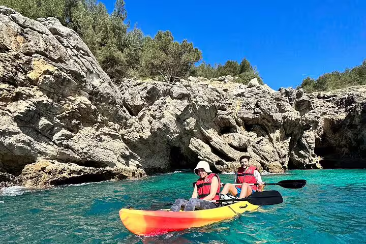 Two kayakers explore the crystal-clear waters and rugged cliffs of Arrábida Natural Park in Sesimbra under a bright blue sky.