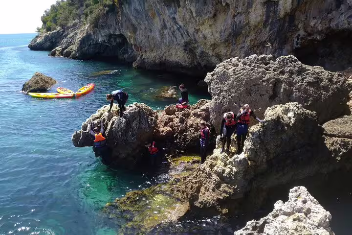 Adventurers coasteering on rocky cliffs in Lisbon with kayaks floating in the clear blue water nearby.
