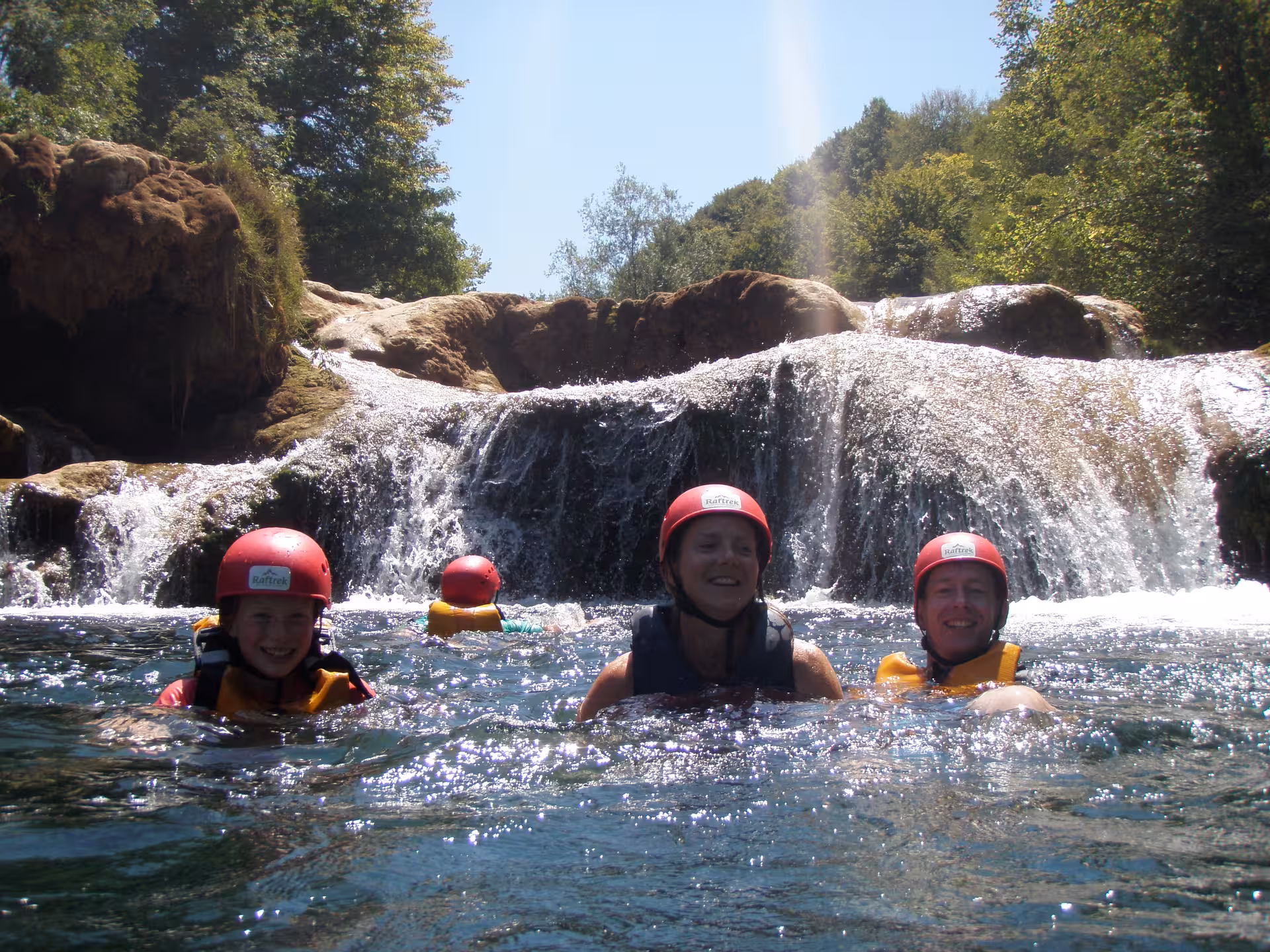 Smiling group wearing helmets enjoying swimming near a waterfall in Mreznica Canyon, ideal for family adventure.