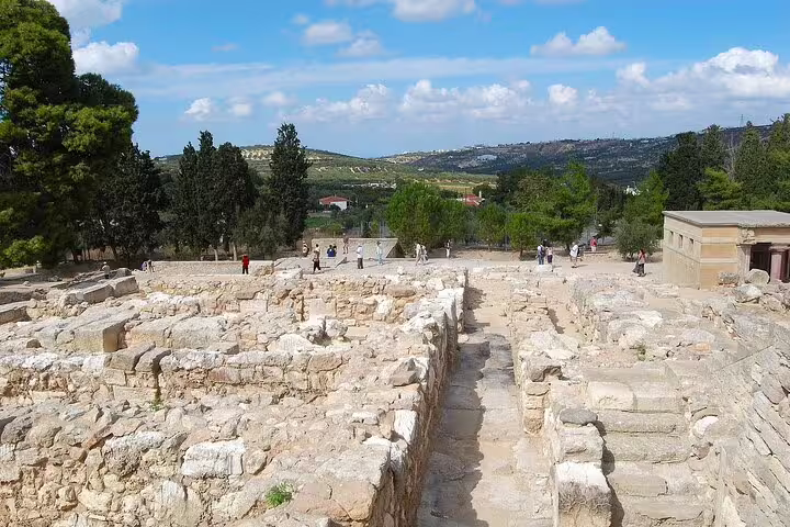Visitors explore the ancient ruins of Knossos Palace surrounded by lush greenery and distant hills under a clear blue sky.