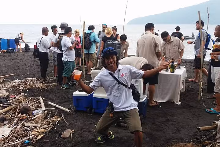 Local guide posing on black-sand beach during Krakatau day tour, with picnic table and island backdrop