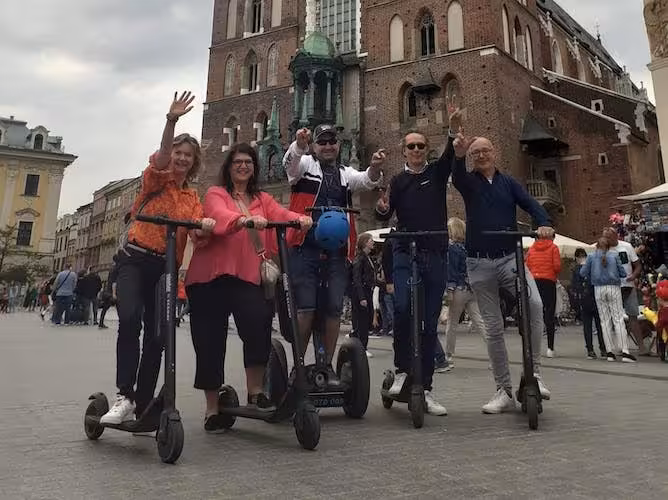Tourists riding e-scooters in Krakow's main square, waving in front of St. Mary's Basilica.