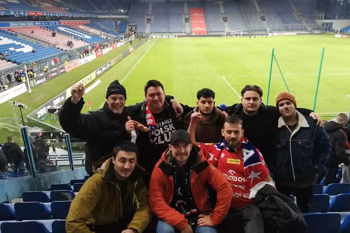 Travelers with local guide at a Krakow football match, posing in the stands with pitch view behind