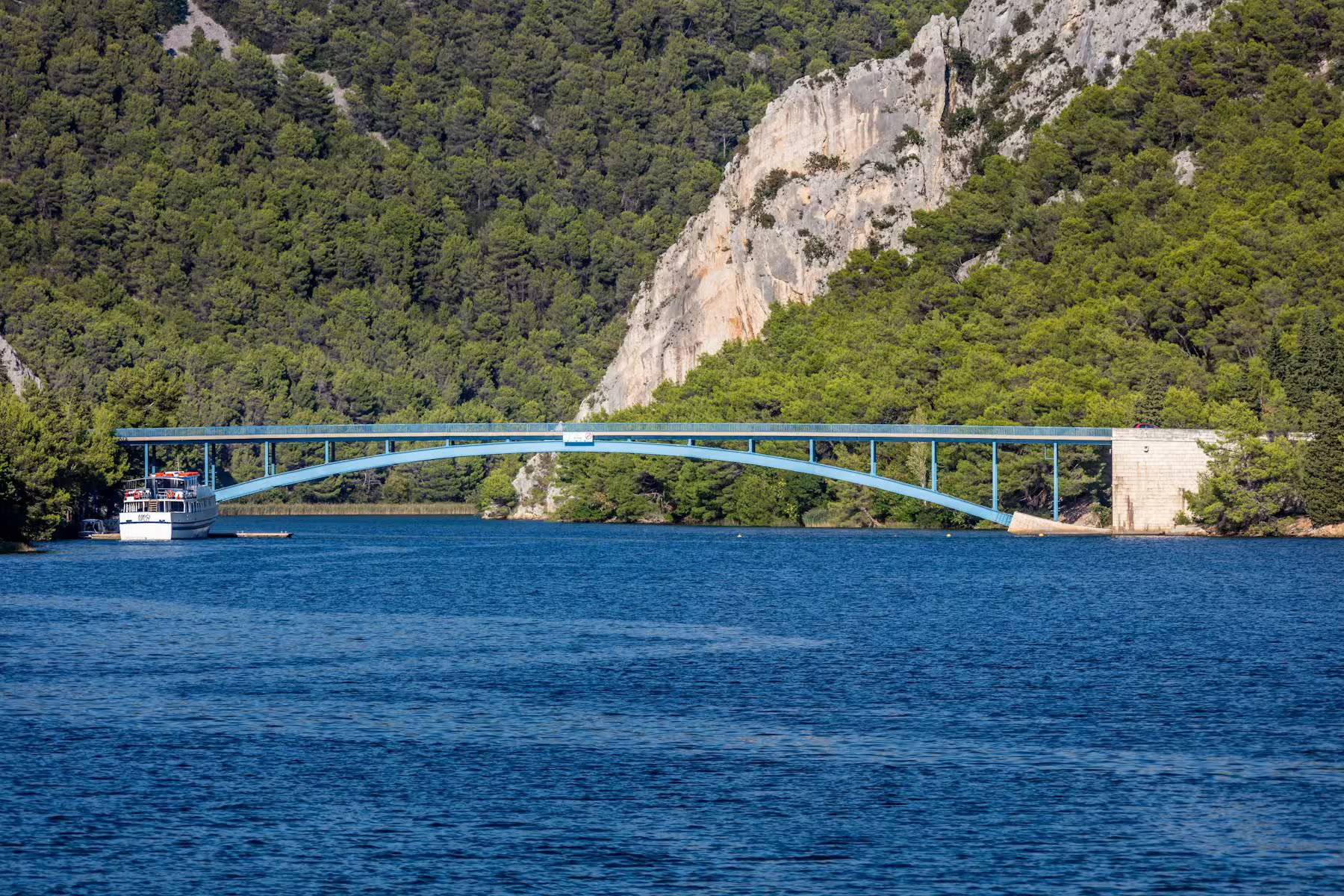 Boat cruising under a blue bridge on the Krka River, part of the Šibenik and Krka National Park tour from Zadar