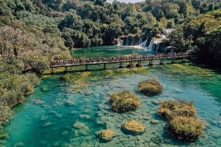 Visitors crossing wooden bridge at Krka Waterfalls above clear emerald water on private tour with wine tasting