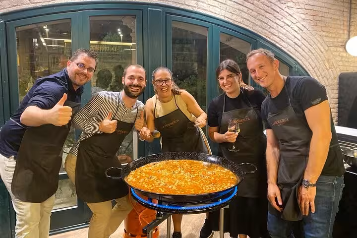 Group enjoying a cooking class at a designer loft, standing around a large pan of freshly prepared paella.