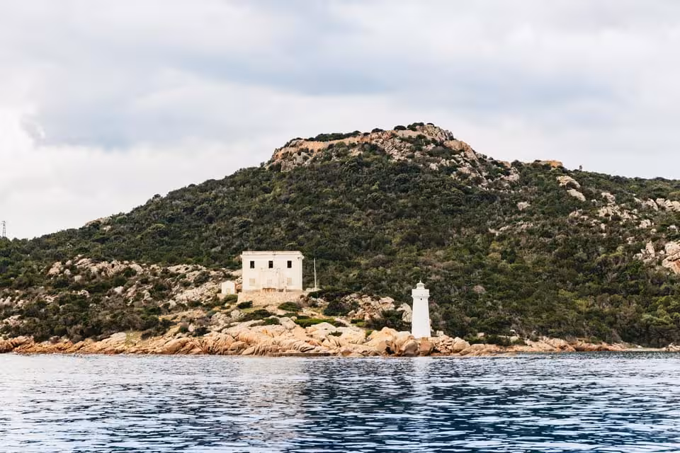 A scenic view of a lighthouse and building on a lush green island in La Maddalena Archipelago, surrounded by calm sea.