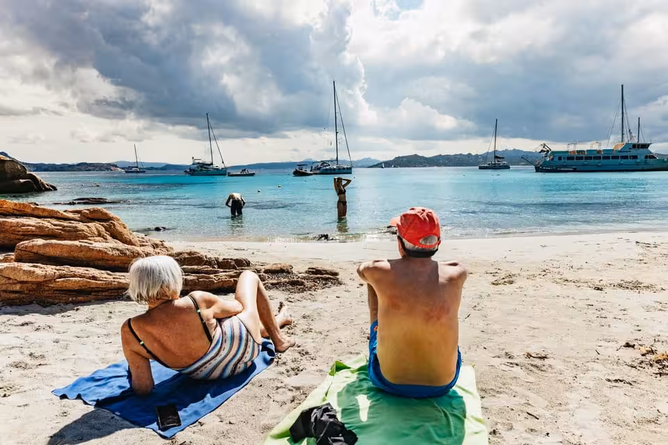 Visitors relax on a sandy beach with yachts in the background in the picturesque La Maddalena Archipelago, Sardinia.