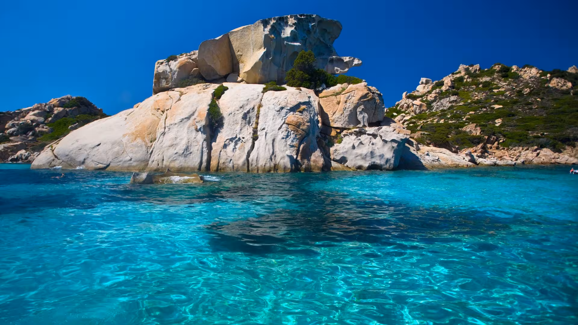 Scenic view of La Maddalena's clear turquoise waters and rugged rock formations on a sunny day.