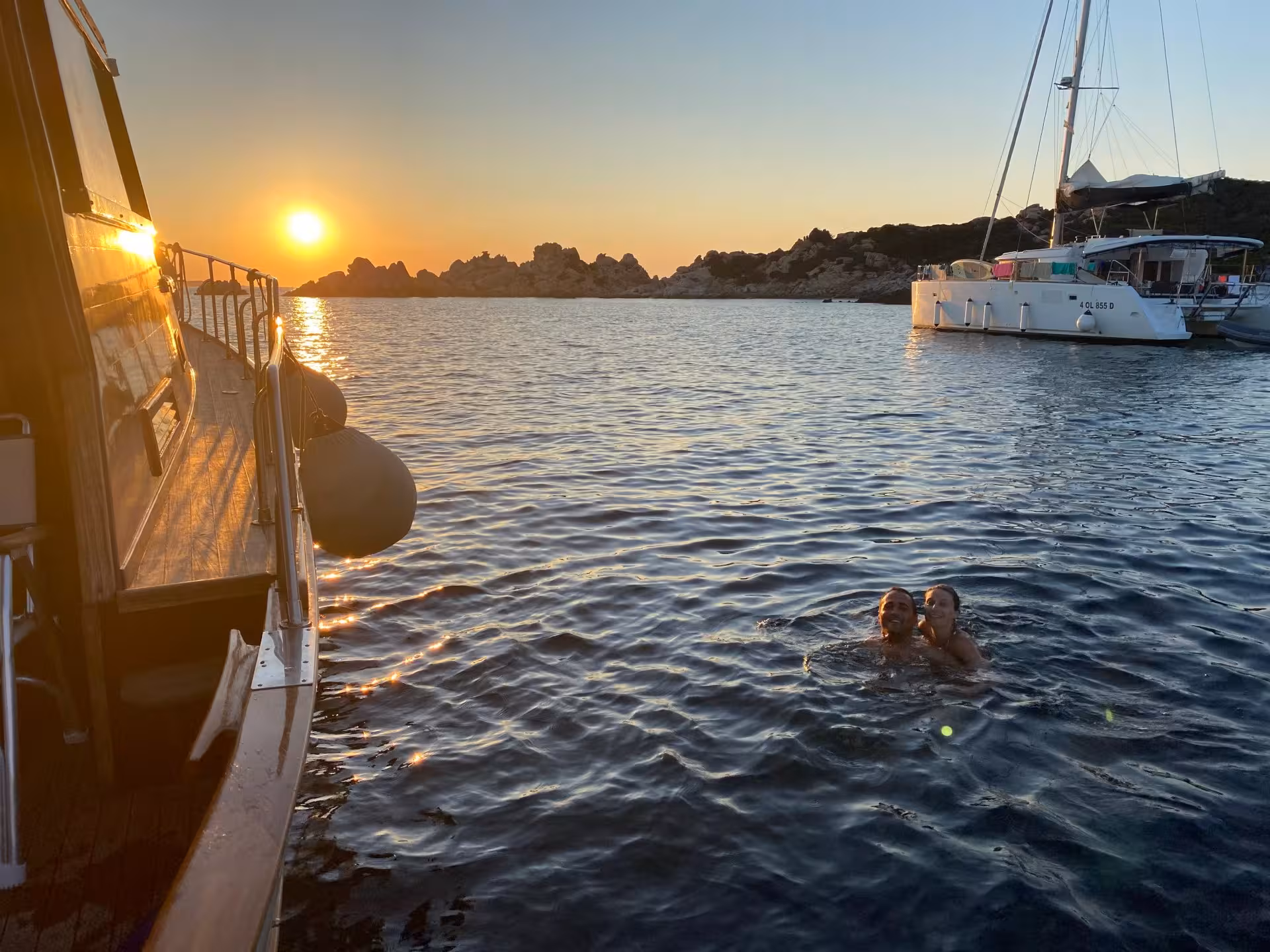 Two people swimming near a boat at sunset in La Maddalena Archipelago, ideal for a private sunset tour adventure.