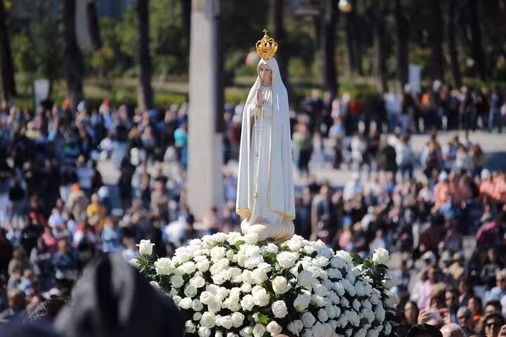 Statue of Our Lady of Fátima adorned with white roses, surrounded by a crowd during a religious event in Portugal.