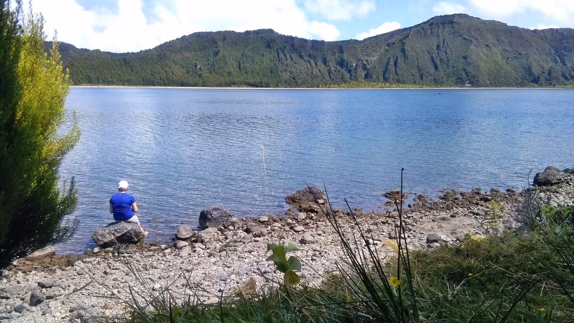 Traveler resting on rocky shore of Lagoa do Fogo during a full-day walking tour hike in São Miguel, Azores