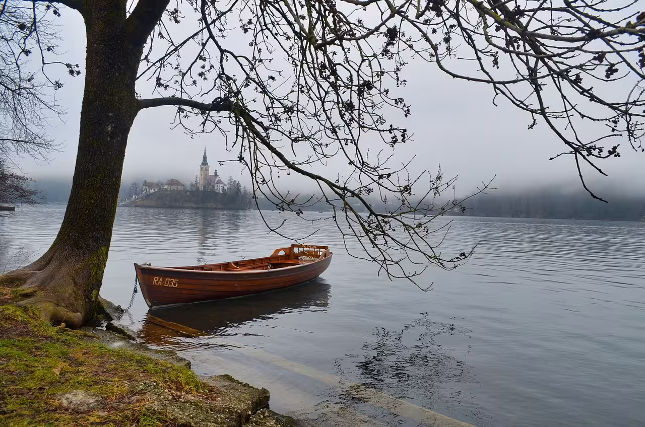 Misty Lake Bled shore with wooden boat and island church in the distance, scenic stop on Ljubljana & Bled tour