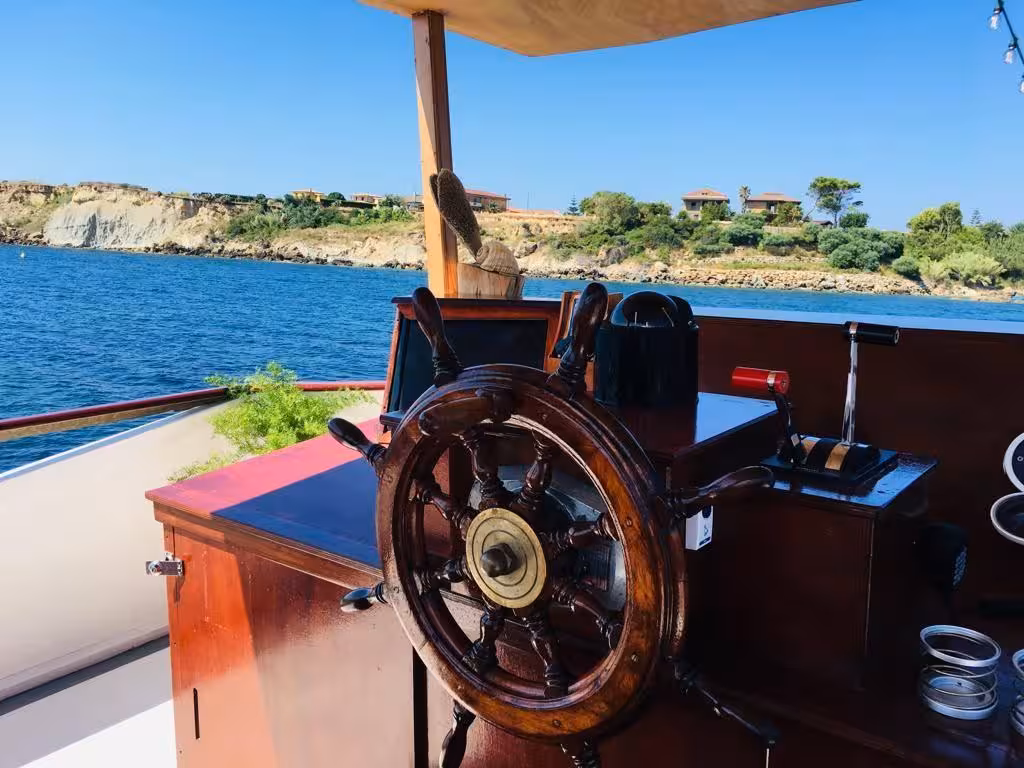 Wooden steering wheel on a boat with scenic view of Capo Rizzuto coastline during Le Castella marine tour.