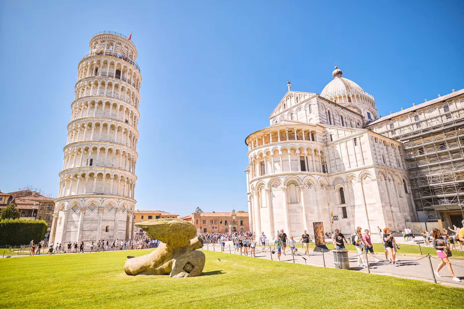 The Leaning Tower of Pisa and Pisa Cathedral with sculpture on green lawn, attracting visitors on a sunny day.