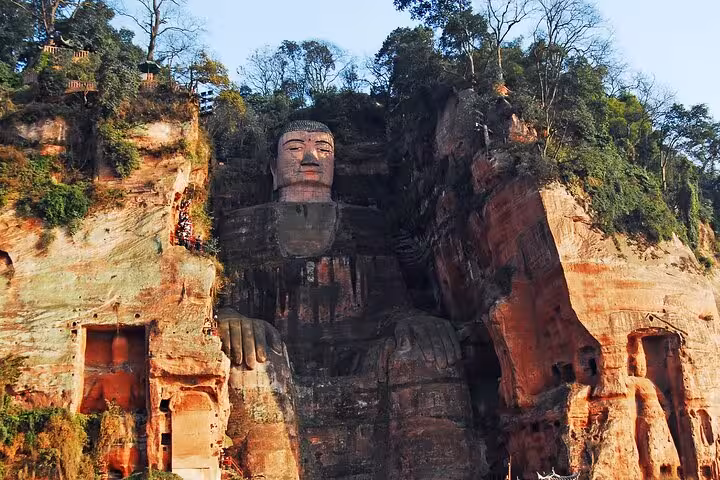 Full view of the Leshan Giant Buddha carved into the red sandstone cliff, framed by lush trees.