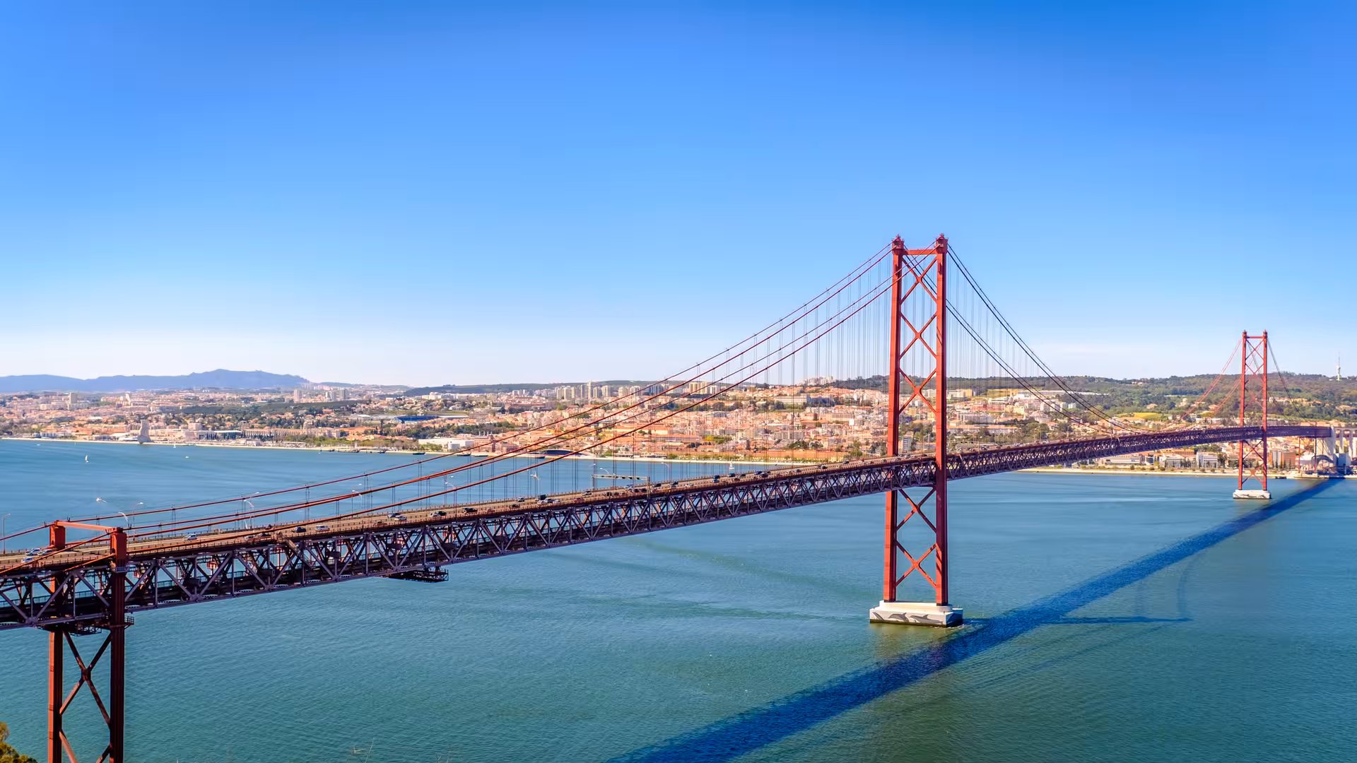 Panoramic view of the iconic 25 de Abril Bridge spanning the Tagus River with Lisbon cityscape in the background.