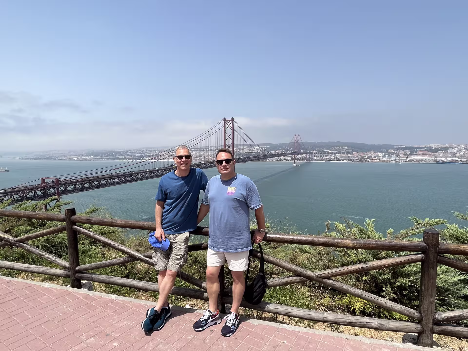 Two tourists enjoying the view of the 25 de Abril Bridge from a Lisbon viewpoint on a clear day.