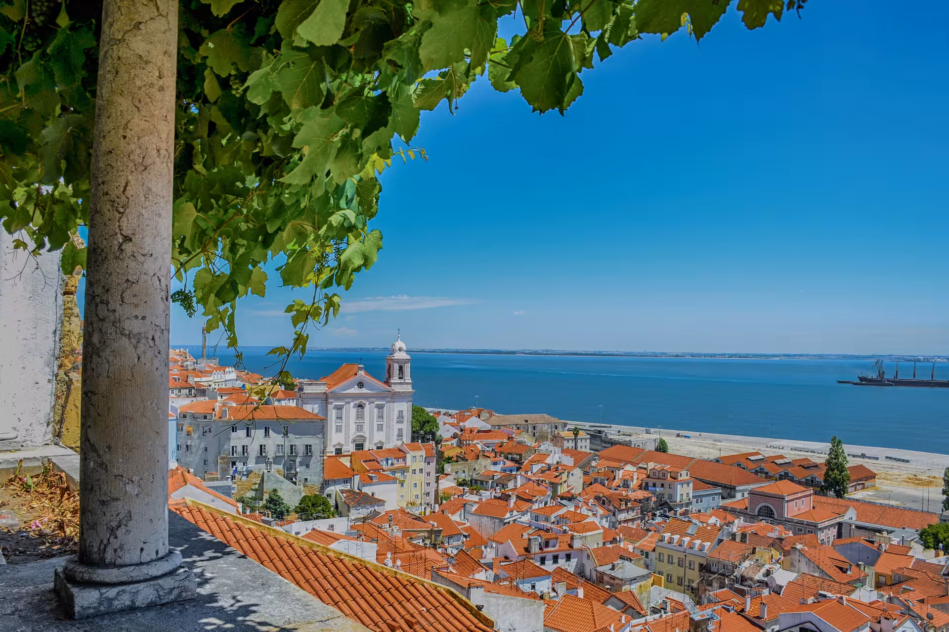 Scenic view of Lisbon's Alfama district with red rooftops and the Tagus River under a bright blue sky.