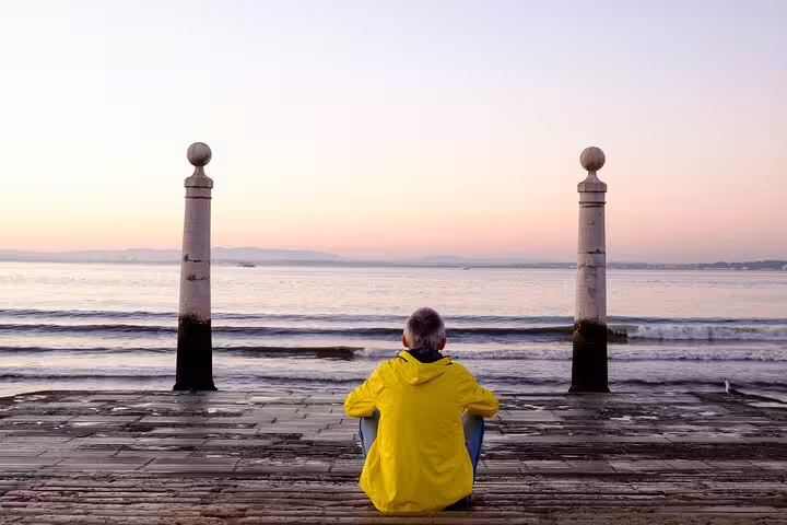 A person in a yellow jacket sits between two pillars, admiring a serene Lisbon waterfront during a private bike tour at sunrise.