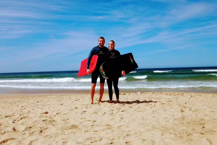 Two people enjoy a sunny bodyboarding adventure on a scenic Lisbon beach, holding boards against a backdrop of blue skies and waves.