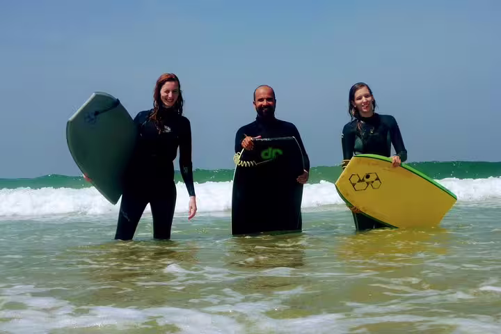 Three people in wetsuits holding bodyboards stand in the ocean, ready for an exciting Lisbon bodyboarding adventure.