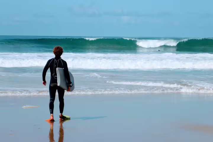 Person in wetsuit holding a bodyboard faces ocean waves on a sunny beach, ready for a Lisbon bodyboarding adventure.