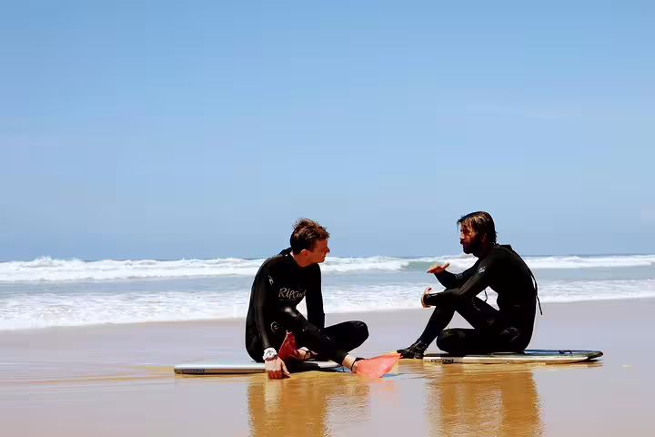 Two bodyboarders in wetsuits sit on surfboards discussing techniques on a sunny Lisbon beach, with waves in the background.