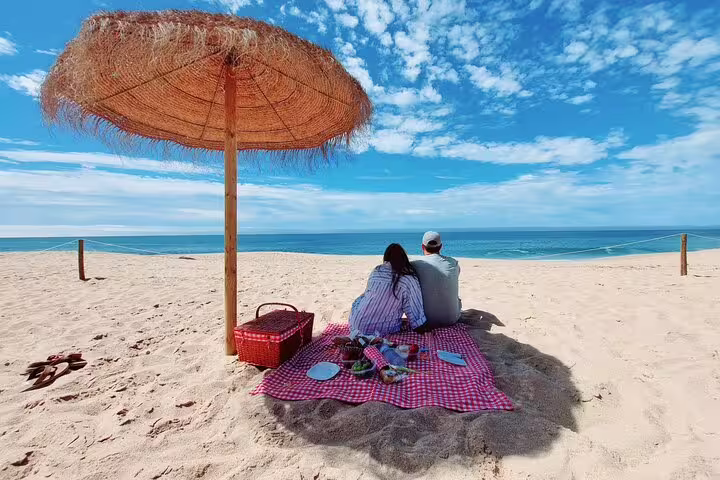 Couple enjoying a beach picnic under a straw umbrella on the Private 4X4 Jeep Tour in Arrábida, Sesimbra-Lisbon.