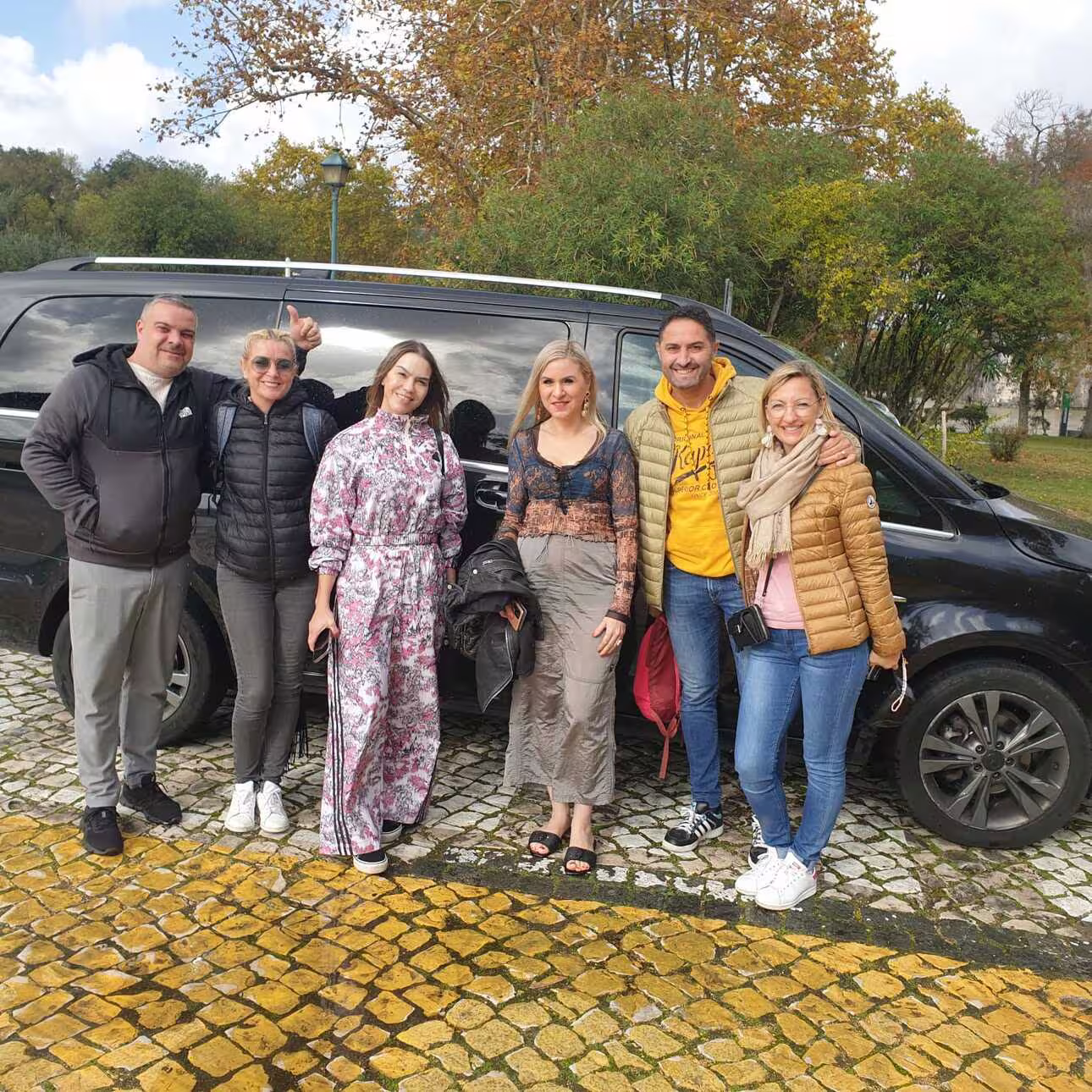 Group of tourists smiling in front of a black van on a Lisbon to Fátima tour with autumn foliage backdrop.