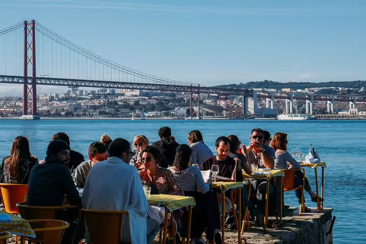 Visitors enjoy a scenic food and drink experience by the water with views of Lisbon's iconic 25 de Abril Bridge on a sunny day.