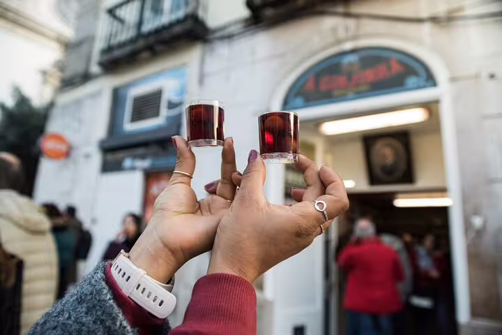 Two hands holding traditional cherry liqueur shots outside a local Lisbon bar, showcasing authentic Portuguese experiences.