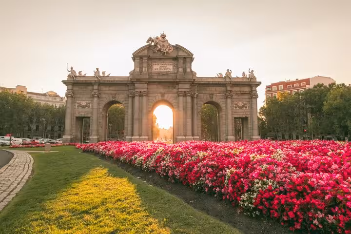Sunset view of Puerta de Alcalá surrounded by vibrant flowers, highlighting a scenic stop on the Lisbon to Madrid transfer.