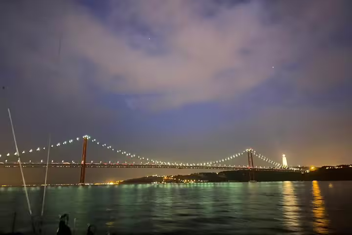 Nighttime view of Lisbon's 25 de Abril Bridge glowing with lights, reflecting beautifully on the Tagus River.