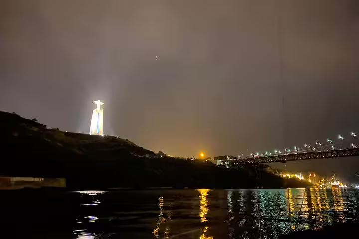 Illuminated Christ the King statue and 25 de Abril Bridge view from a nighttime Lisbon sailing tour on the Tagus River.