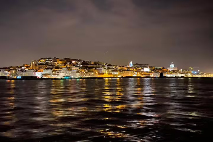 Lisbon skyline at night from a sailing perspective, highlighting historic buildings and their reflections on the water.