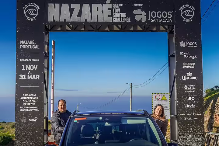 Tourists stand by a car under the Nazare Surf Challenge sign with ocean views on a Lisbon to Porto private tour.