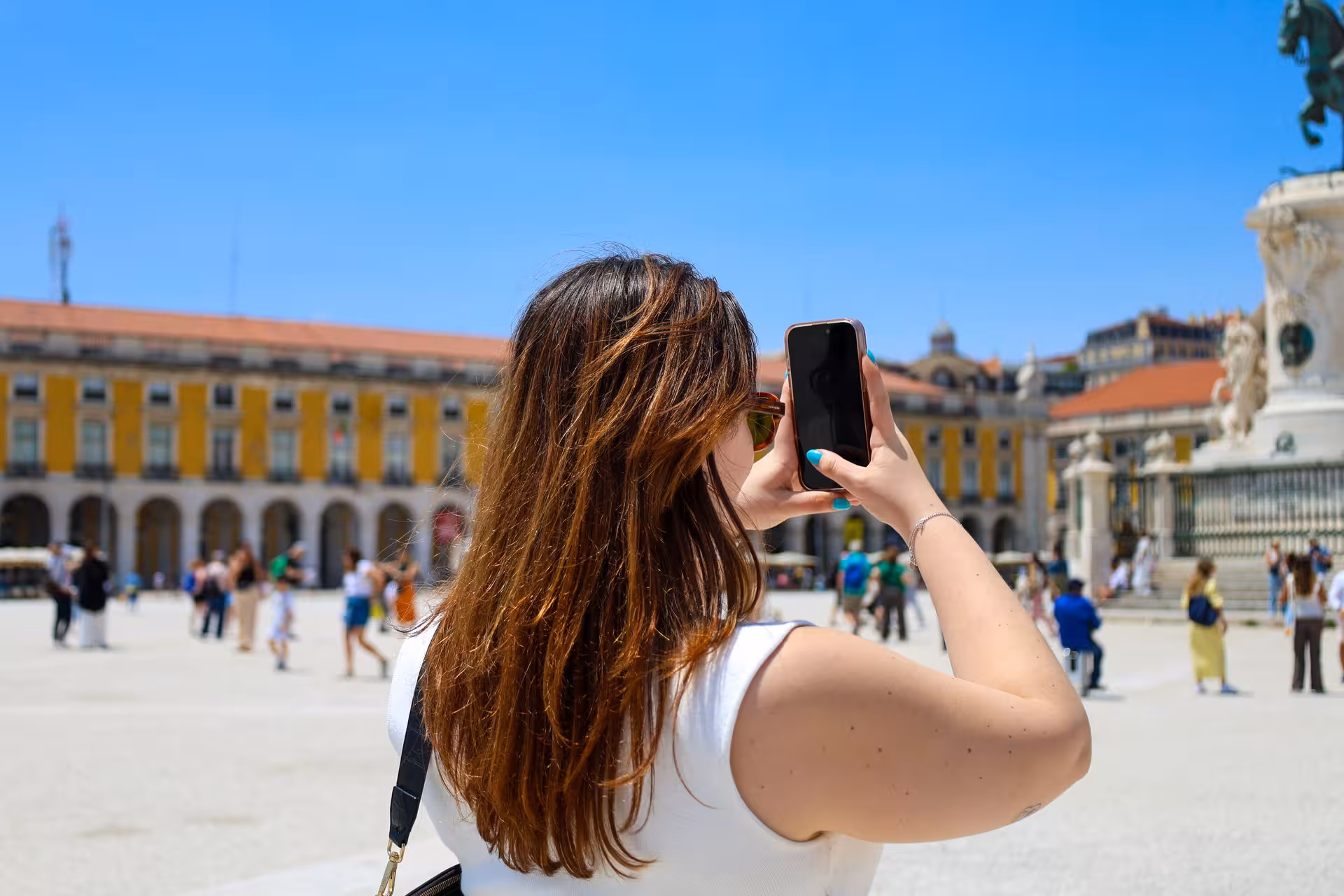 Tourist capturing the vibrant atmosphere of Lisbon's Praça do Comércio with a smartphone, highlighting city exploration.