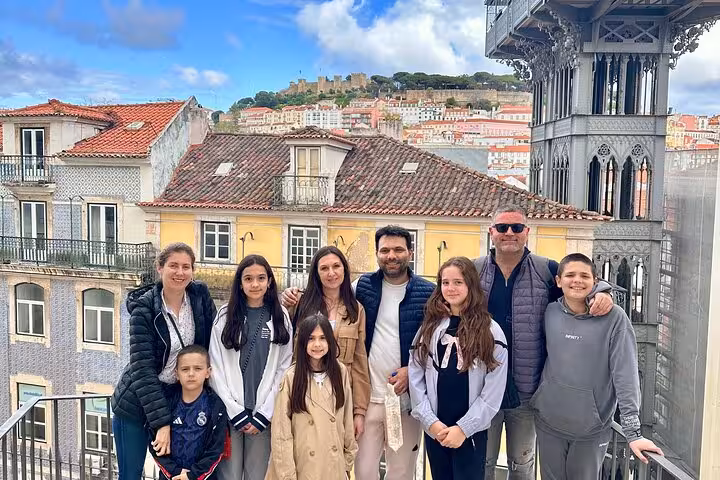 Family posing at Lisbon's Santa Justa Lift with a view of the city's historic architecture and São Jorge Castle.