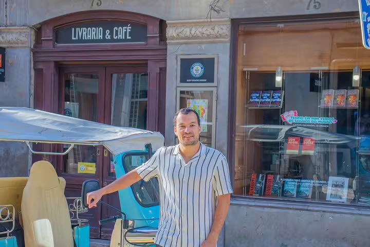 Man standing beside a TukTuk in front of Lisbon café, ready for a private tour with local guide, showcasing city exploration.