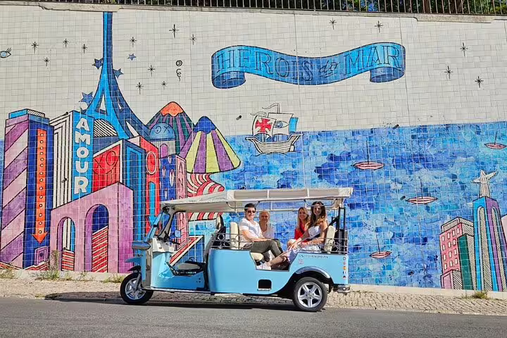 Blue tuk-tuk with happy tourists on a vibrant Lisbon street, showcasing colorful murals during a private guided tour.