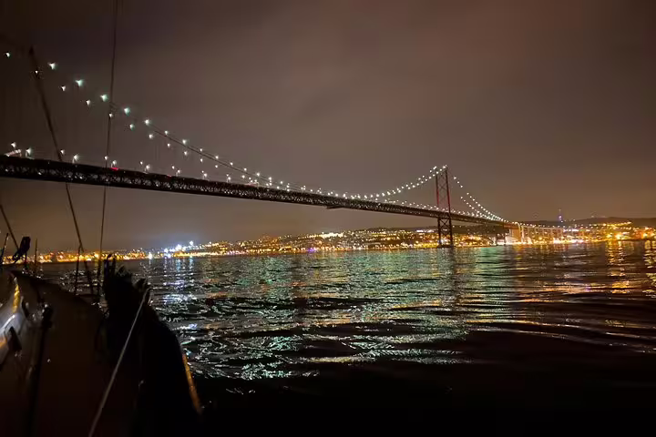 Sailing under Lisbon's illuminated 25 de Abril Bridge at night, capturing city lights reflecting on the Tagus River.