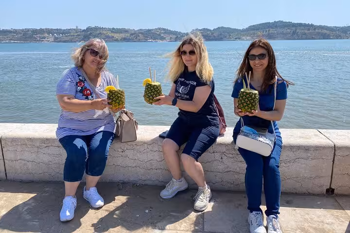 Three women enjoy pineapple drinks by the river, highlighting the Lisbon to Sintra guided tour's local charm.
