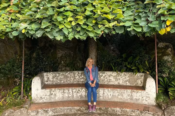 Smiling woman seated on a stone bench under leafy foliage in the gardens of Sintra's Regaleira estate during a guided tour.