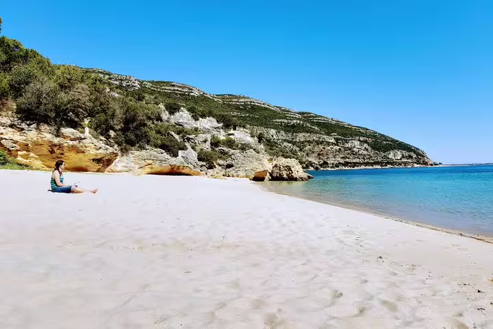 Serene beach view on Lisbon's southern coast, featuring golden sands, clear blue waters, and lush green hills under a bright sky.
