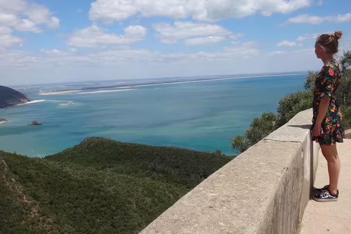 A tourist enjoys the stunning coastal view from a scenic overlook on Lisbon's South Sights & Wine tour, highlighting lush greenery and blue waters.