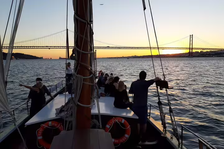 Sunset over Lisbon's bridge as tourists enjoy a serene cruise with wine on a traditional boat.