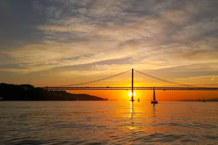 Sunset view of 25 de Abril Bridge from Tagus River on Lisbon sailing cruise, calm waters and sailboats